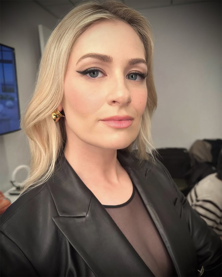 A woman with long blonde hair, wearing a black blazer and gold earrings, looks confidently at the camera in a well-lit indoor setting.
