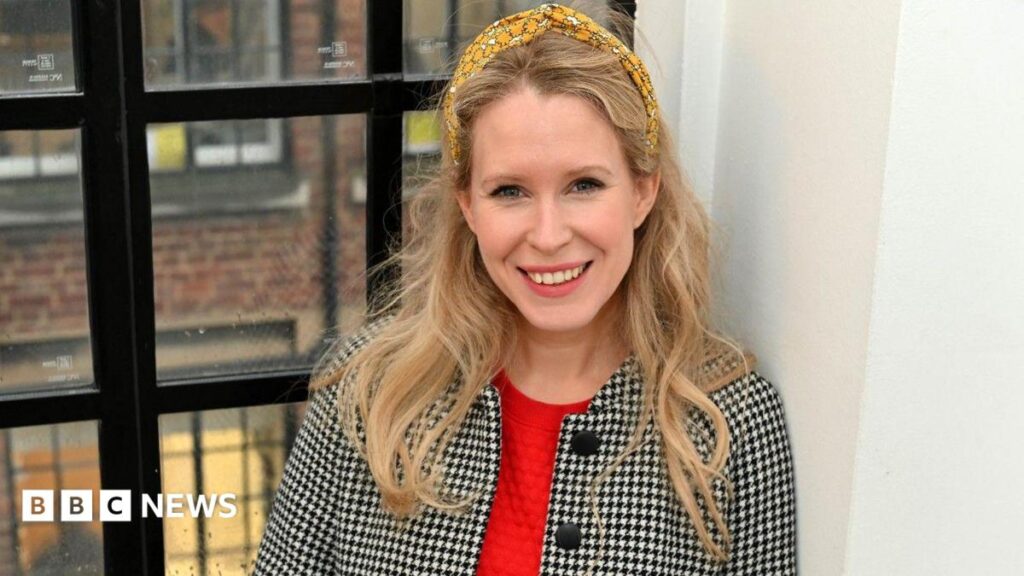 Lucy Beaumont - a woman with long wavy blonde hair wearing a flowery yellow headband - is smiling into the camera and leaning against a wall by a window.  She is wearing a black and white tailored jacket with a red top.