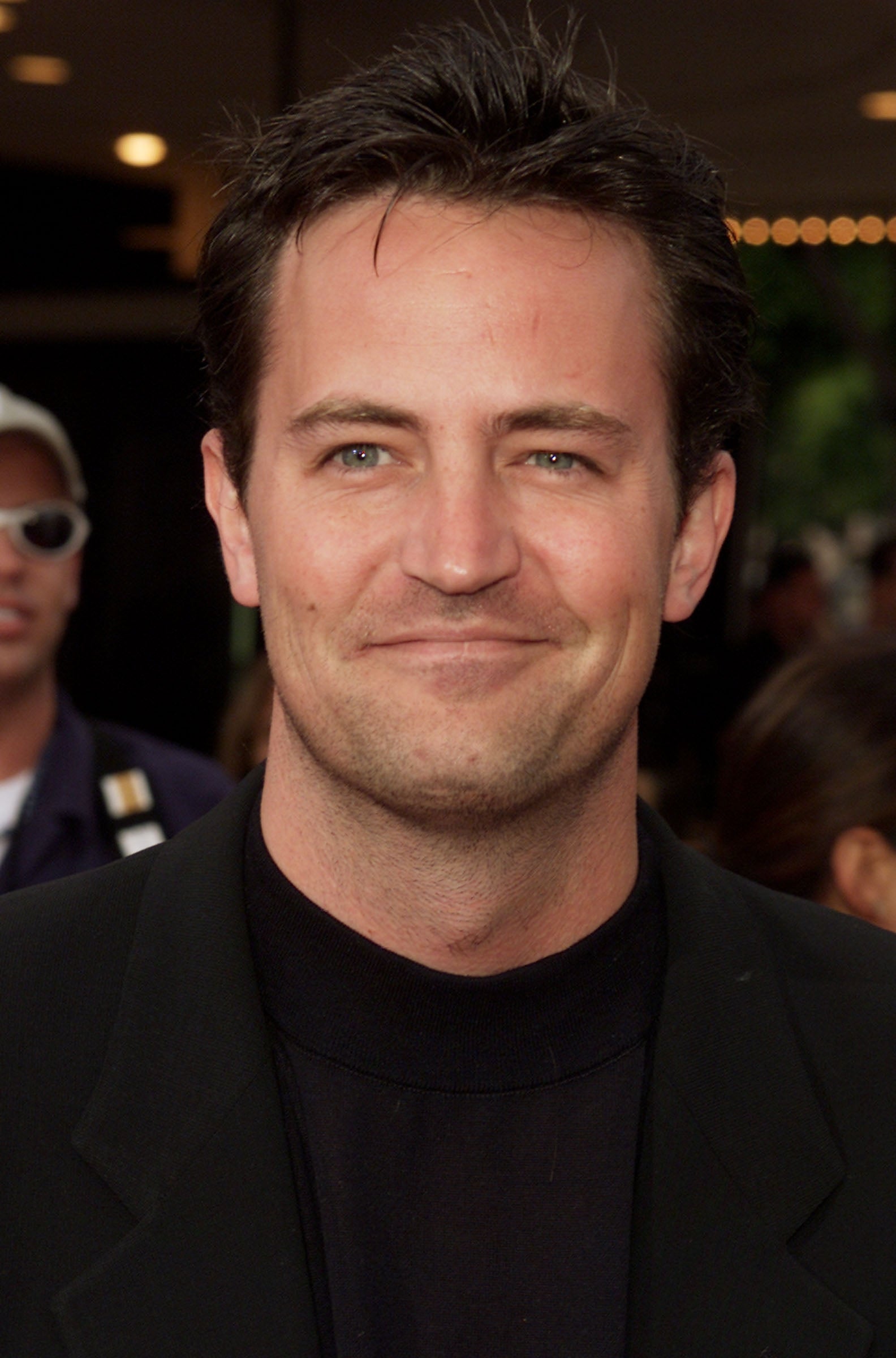 Matthew Perry smiling at an event, wearing a black jacket and crew-neck shirt