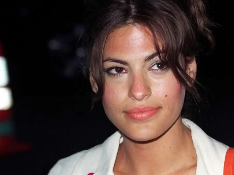 A woman with medium skin tone and brown hair looks slightly to the side, wearing a white top. The background is dark with blurred lights.