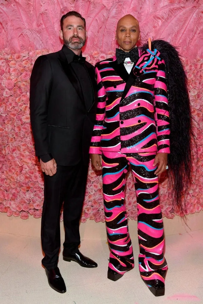 Two individuals on a red carpet. One in a classic black tuxedo, the other in a vibrant patterned suit with bold designs. Pink flower backdrop