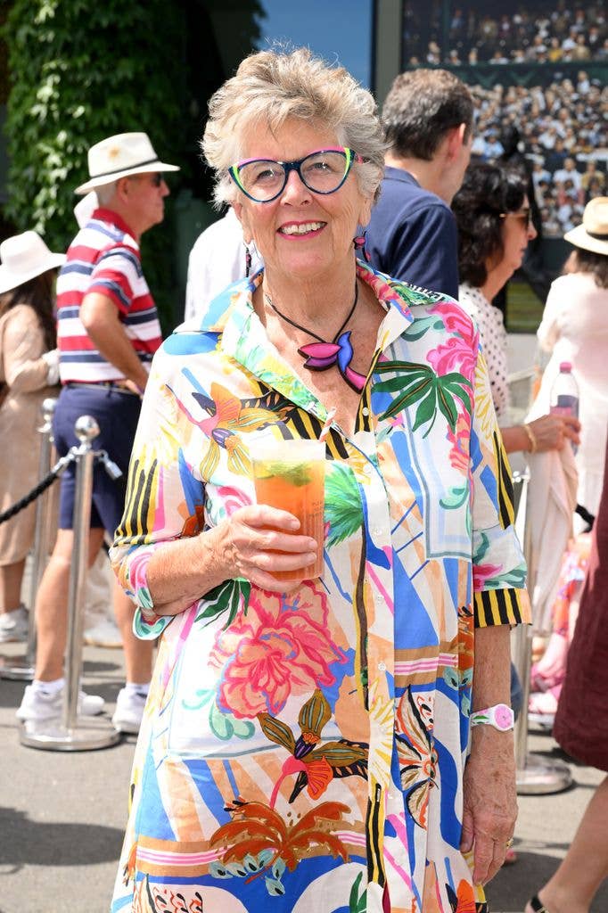 Prue Leith, wearing a colorful shirt and glasses, holds a drink and smiles.