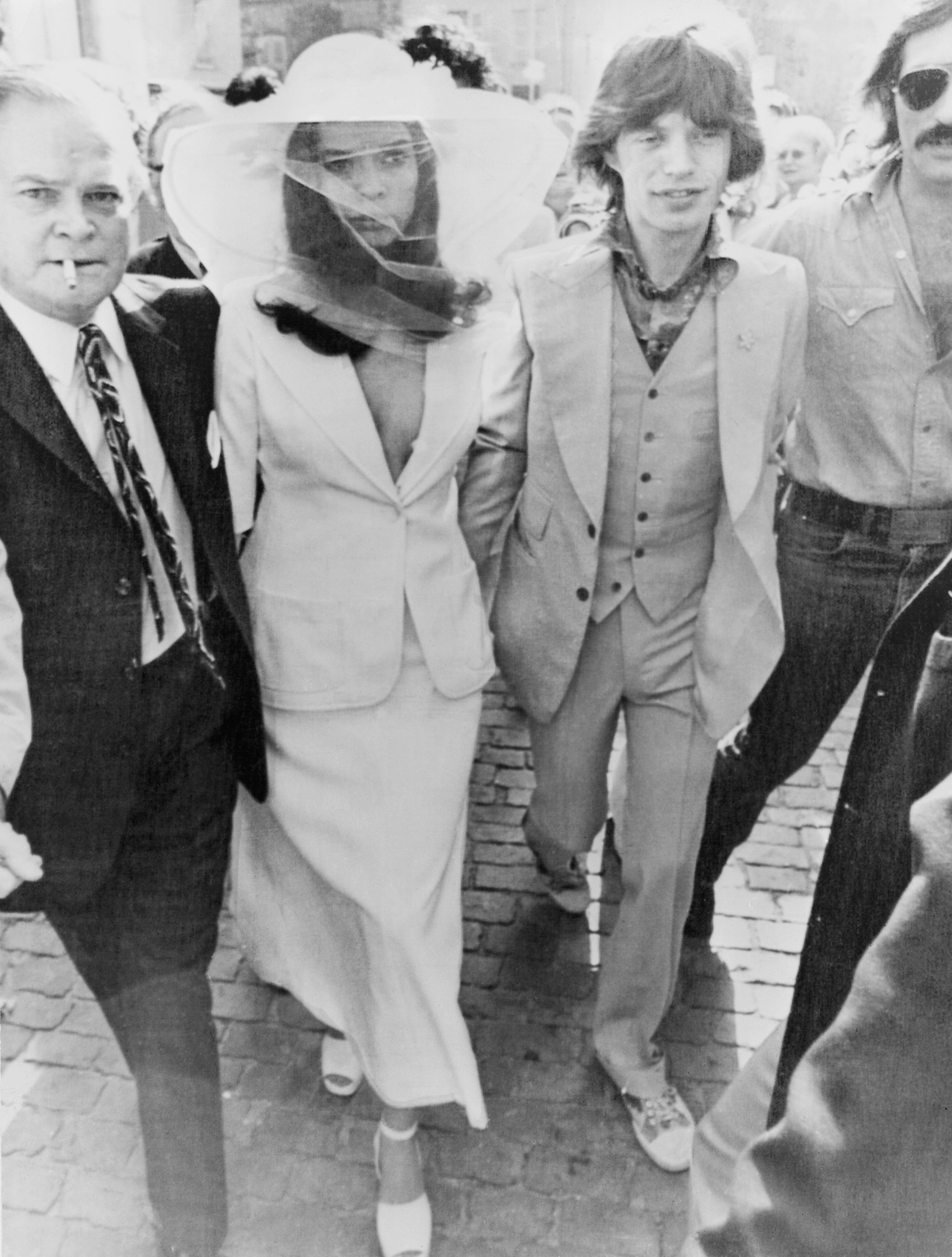 A celebrity couple walks with others. The woman wears a stylish hat with a veil and a chic suit, while the man sports a tailored suit with an open-lapelled shirt