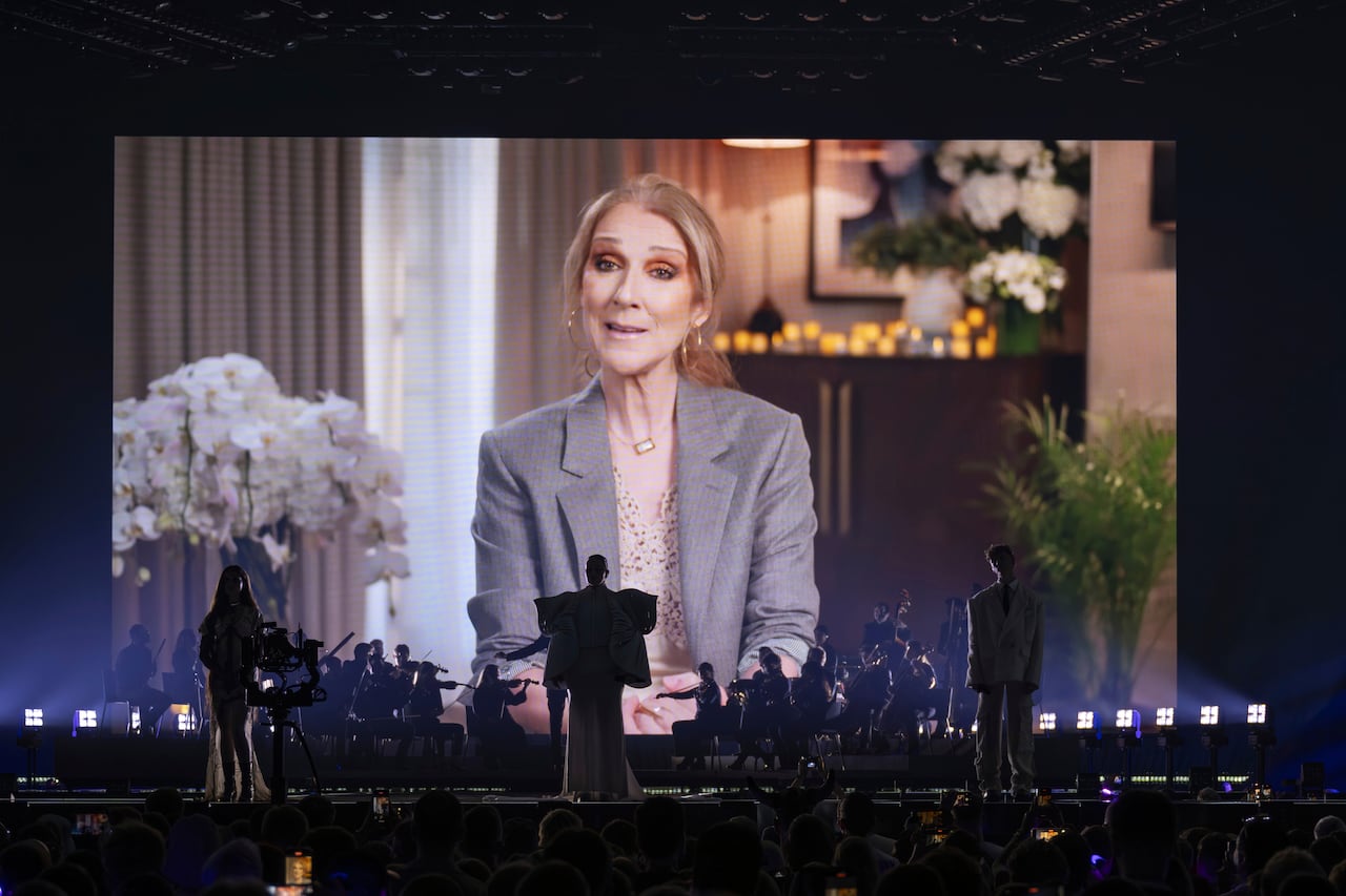 A band is seen on stage in shadow, backlit by Celine Dion appearing from a living room on screen.