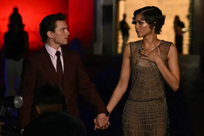 Two individuals holding hands on a red carpet. The woman wears a netted dress; the man is in a suit. They are gazing at each other
