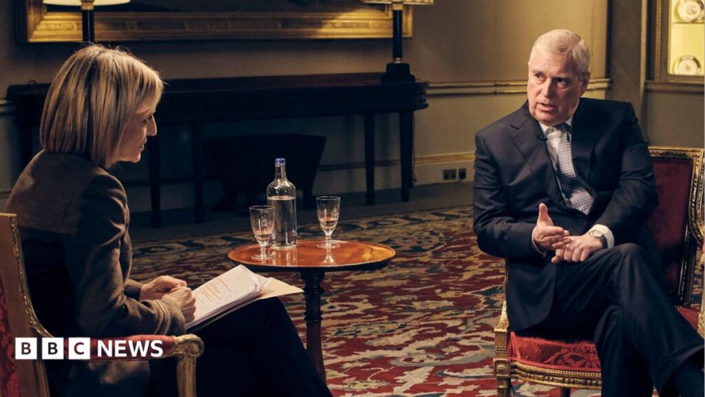 Two people seated opposite each other in upholstered armchairs during a filmed interview in a formal room. A small round table between the chairs holds a glass bottle of water and two drinking glasses. Studio lighting, light stands, microphones and cables are visible around the set. The room has a patterned red-and-cream carpet, decorative wall displays, table lamps, columns and a large framed painting in the background.