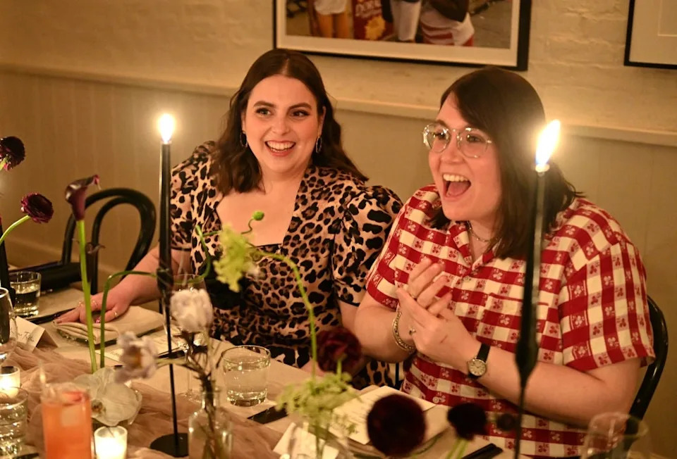 Two people happily sitting at a candlelit dinner table. One wears a leopard print top, the other a red checkered shirt. Decor includes flowers and candles