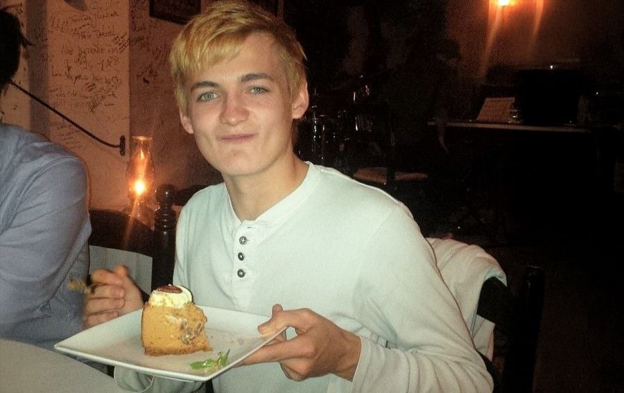 A young person with blond hair, wearing a white shirt, smiles while holding a plate with a slice of cake and a fork, sitting in a dimly lit restaurant.
