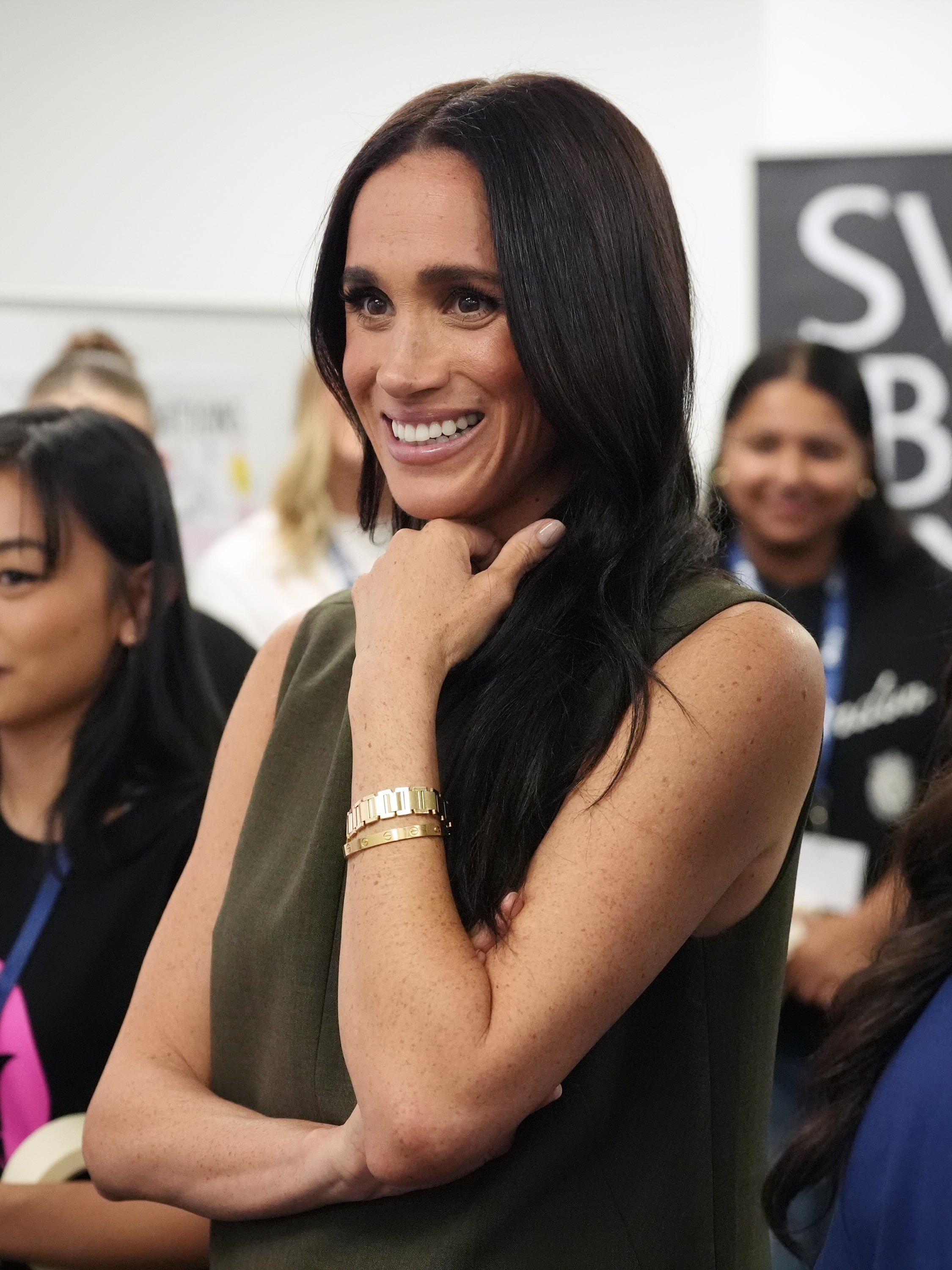 Woman smiling at an event, wearing a sleeveless dress and a gold bracelet. Other attendees are in the background