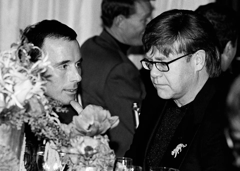 Two people are having a conversation at a formal event. One is wearing glasses and a patterned suit jacket. Flowers are visible on the table