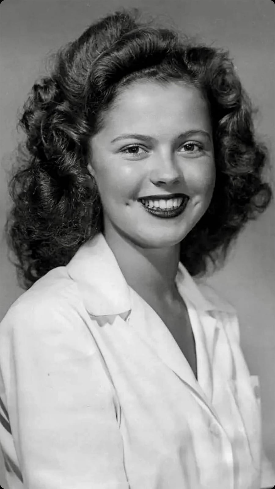 A young woman with wavy dark hair smiles at the camera. She is wearing a white collared shirt. The photo is in black and white, with a plain background.
