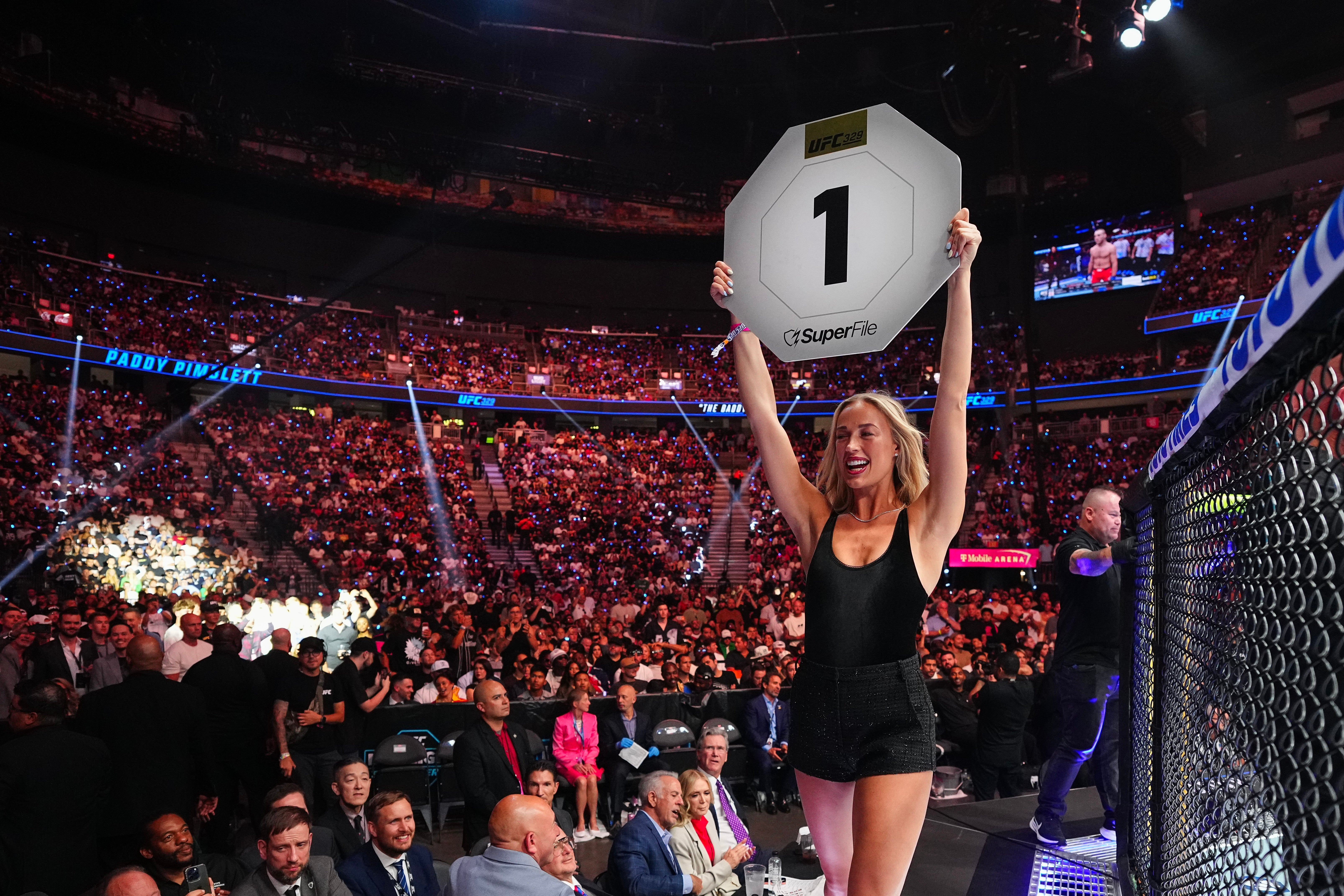 Sophie Cunningham as a ring girl in an outfit holds a sign with the number 1 in a lively, crowded arena at a UFC fight
