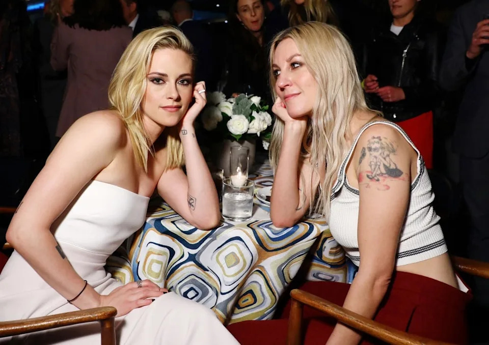 Two individuals sit at a table at an event, each with their chin resting on their hand. One wears a strapless dress, the other a tank top