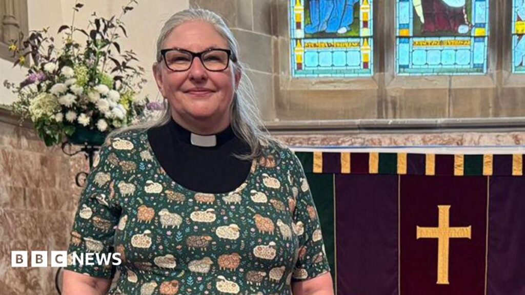 ‘Miracle’ hidden hoard of gold coins saves Melling church Rev Jane Lee with long grey hair wearing black glasses and a black and white clerical collar with a green cardigan over the top with patterns of sheep and flowers on it. She is standing inside St Wilfrid's Church in Melling near the altar.