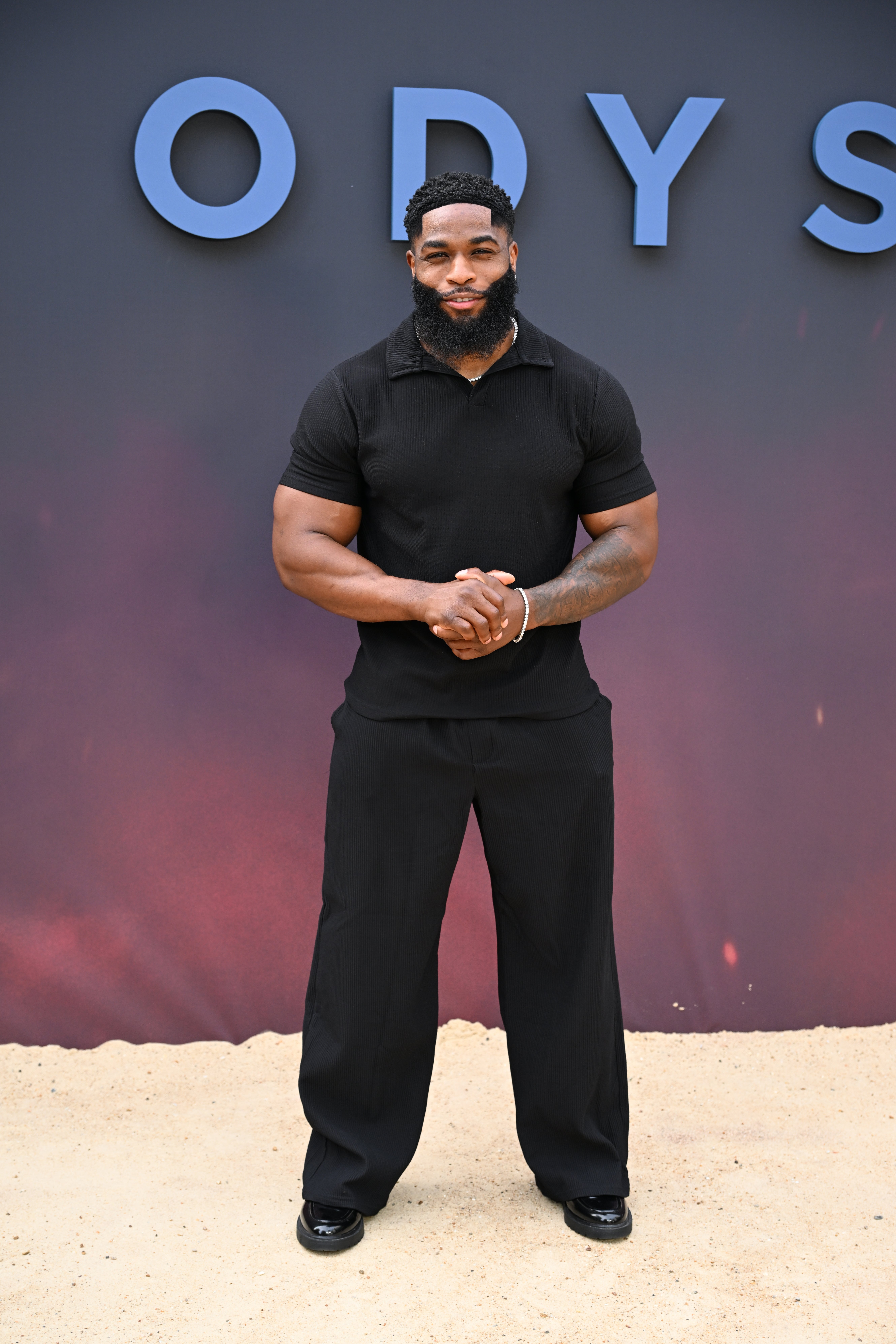 Person on red carpet in casual black outfit, with fitted shirt and wide-leg pants, standing in front of a backdrop with large letters