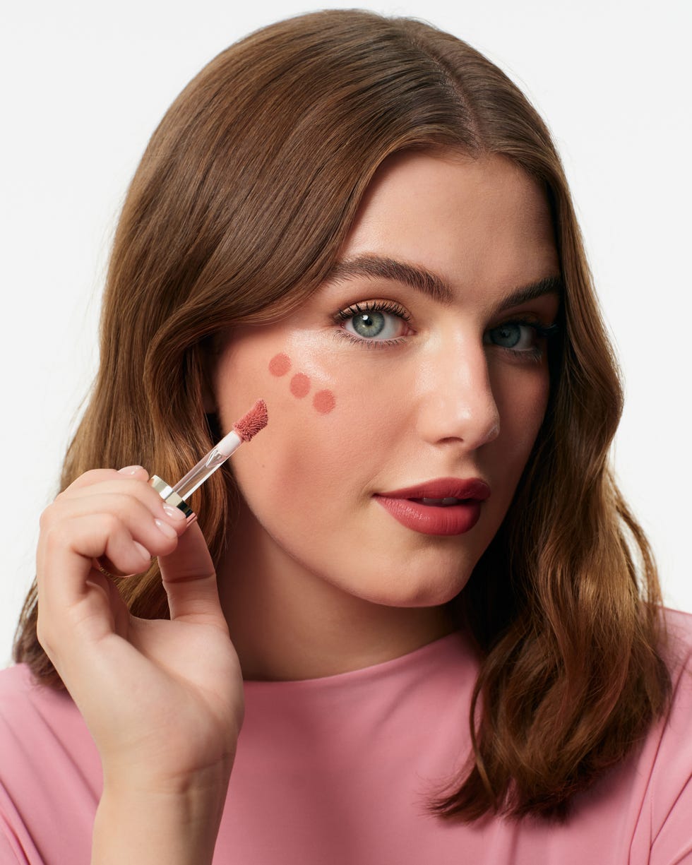 A woman applying blush to her cheek with a brush while wearing a pink shirt. A woman applying blush to her cheek with a brush while wearing a pink shirt.