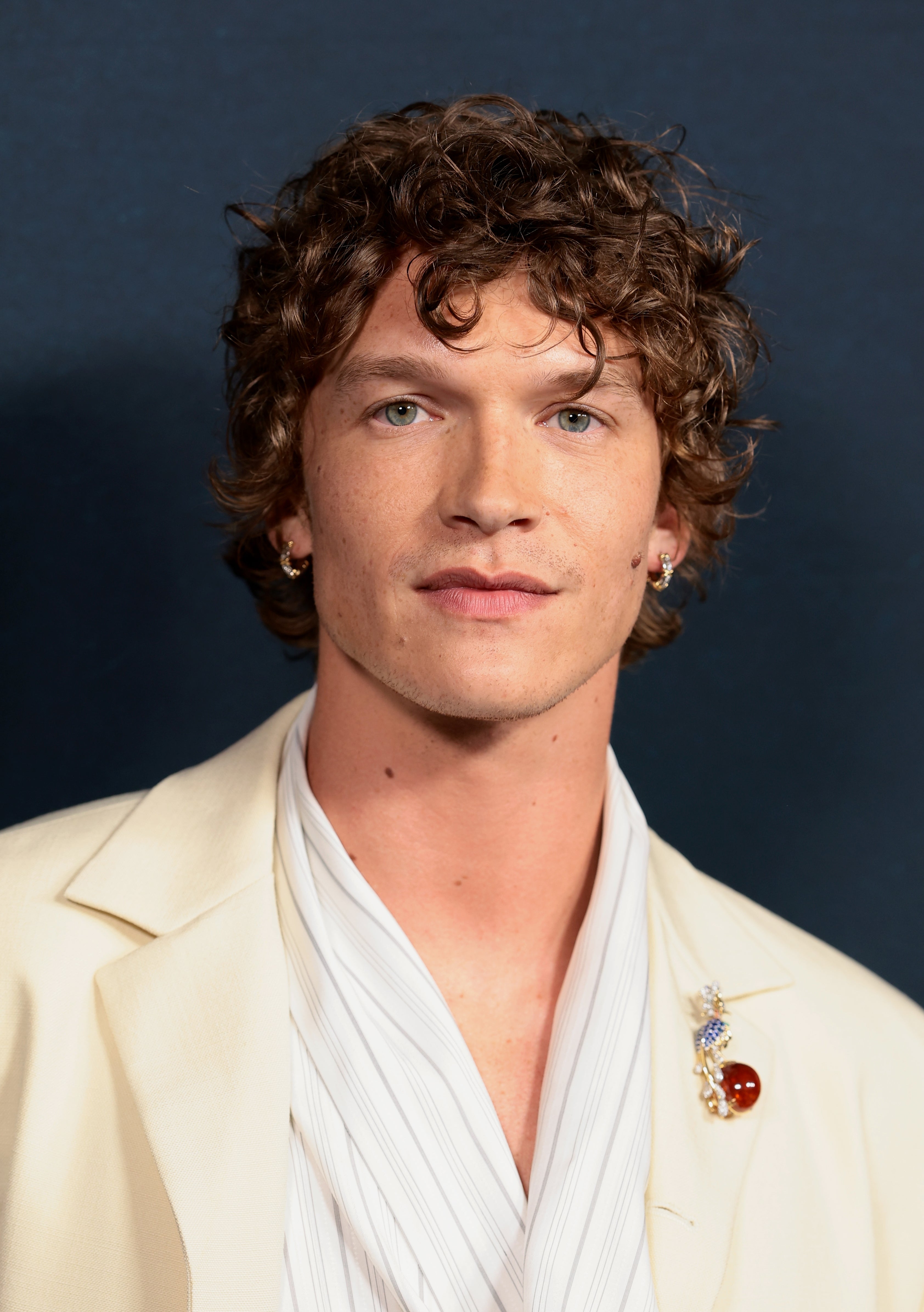 Person in a formal cream-colored suit and white shirt with curly hair, looking at the camera