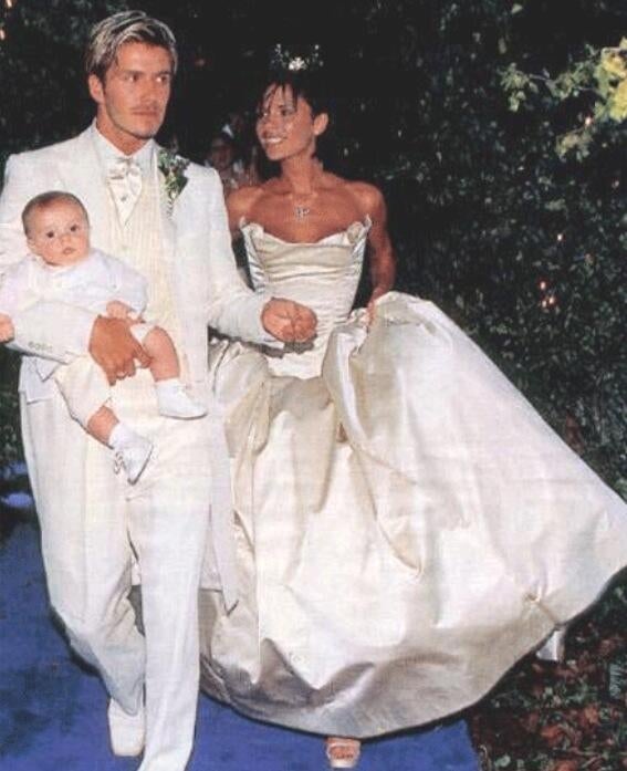 A man in a white suit holds a baby, walking beside a woman in a strapless gown and tiara on what appears to be a wedding day