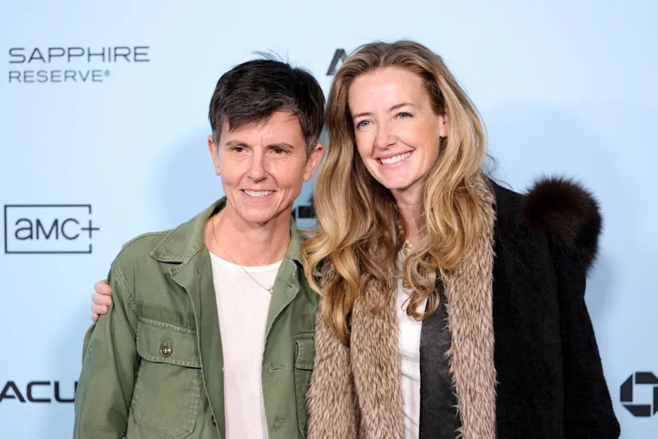 Two people smiling, one wearing a green jacket and white shirt, the other in a coat with a scarf. They are on a press event backdrop