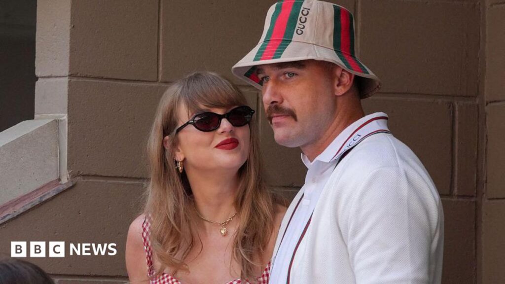 Taylor Swift, wearing a red gingham top, looking up at Travis Kelce, wearing a striped Gucci bucket hat