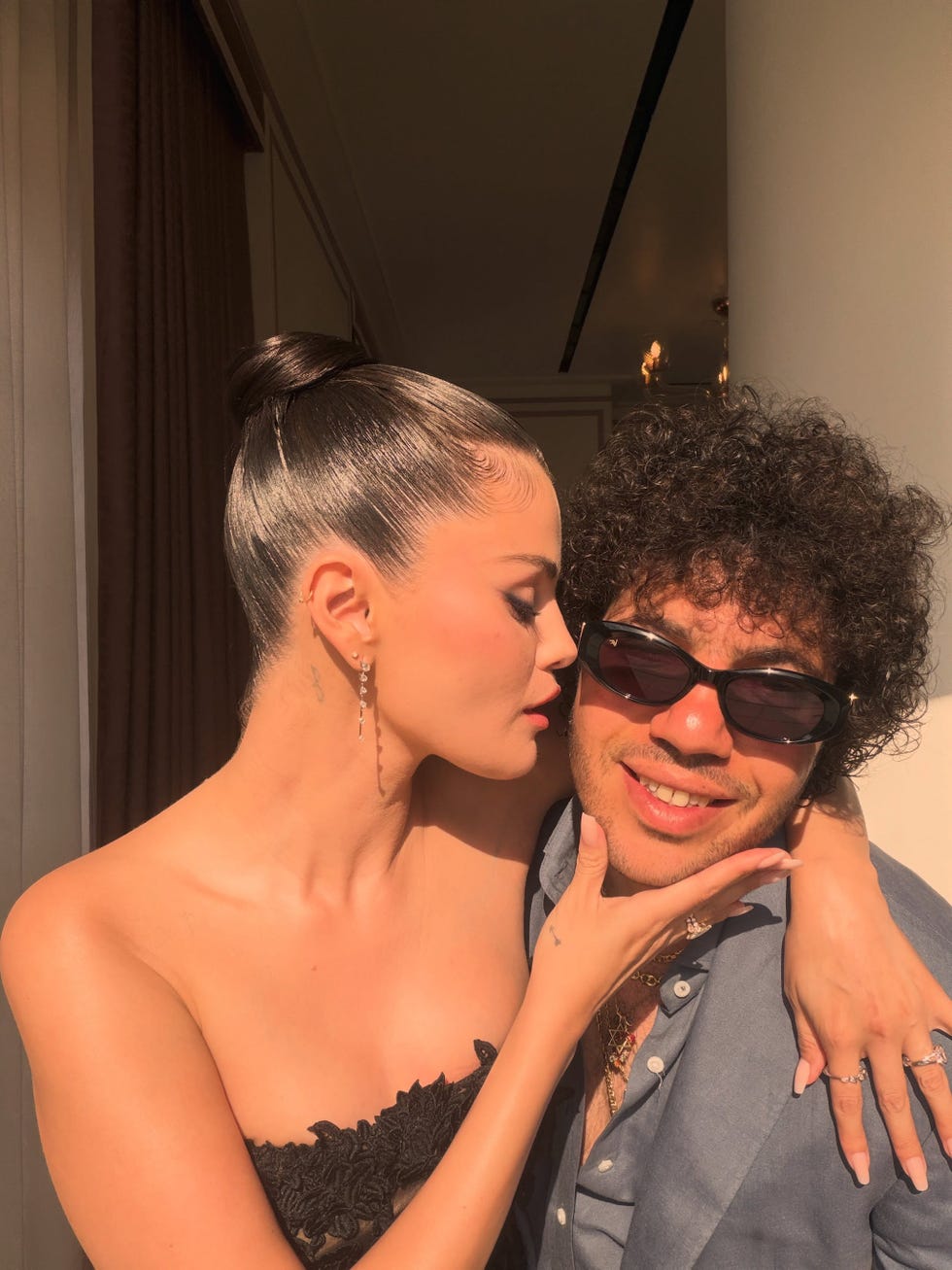 A woman in a black dress leans in to kiss a man wearing sunglasses, both smiling against a bright background. A woman in a black dress leans in to kiss a man wearing sunglasses, both smiling against a bright background.