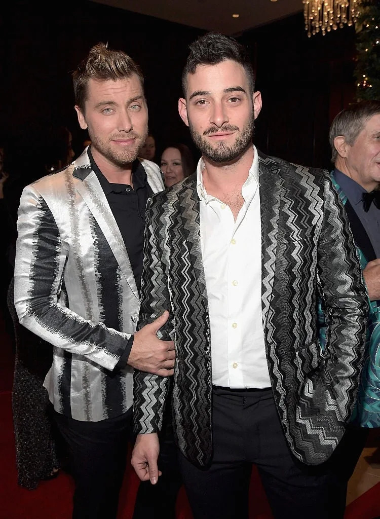 Two individuals at a formal event in matching zigzag patterned blazers, one with a hand on the other's back, smiling at the camera