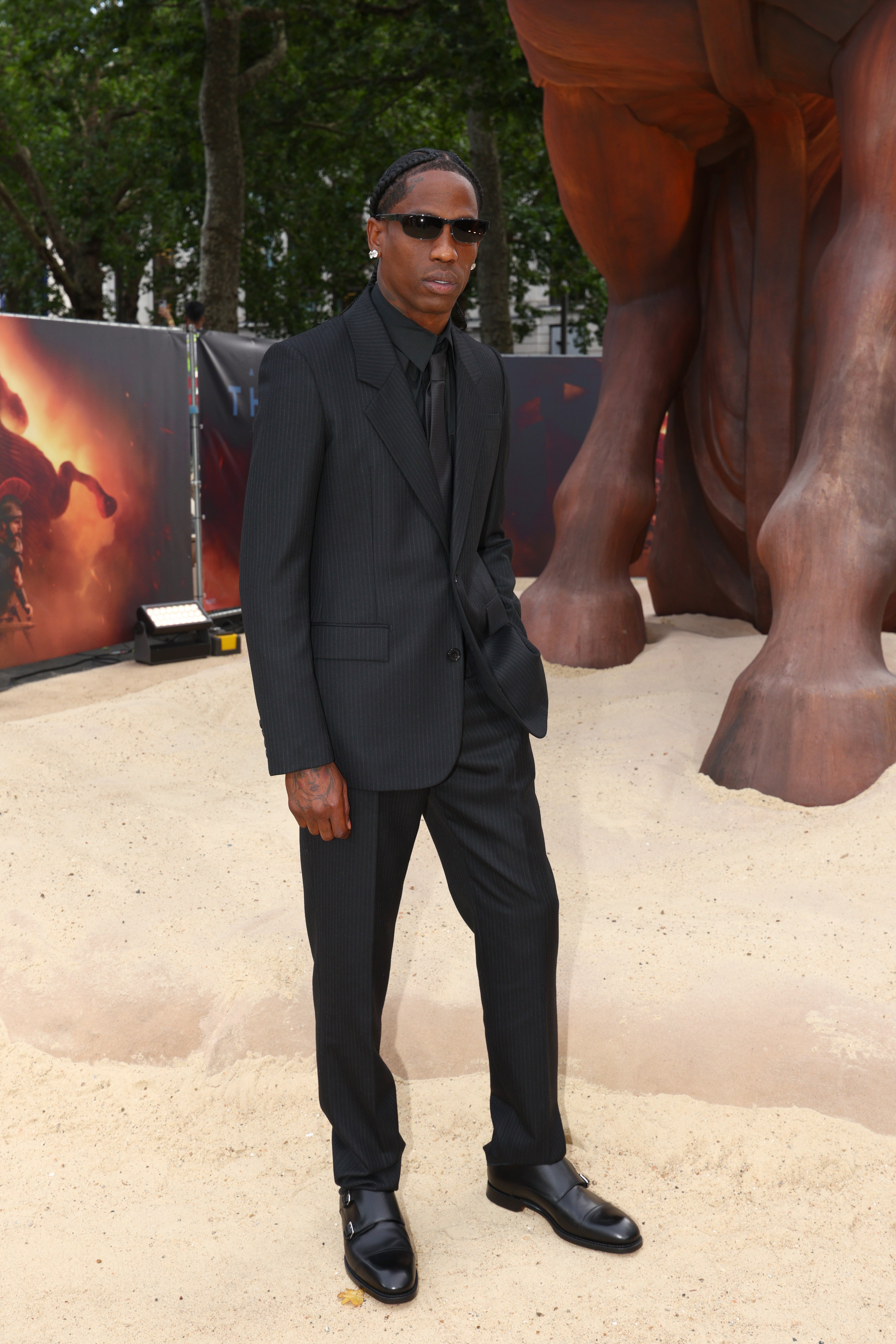 A person in a dark pinstripe suit with sunglasses stands on a sandy surface at a public event, posing stylishly