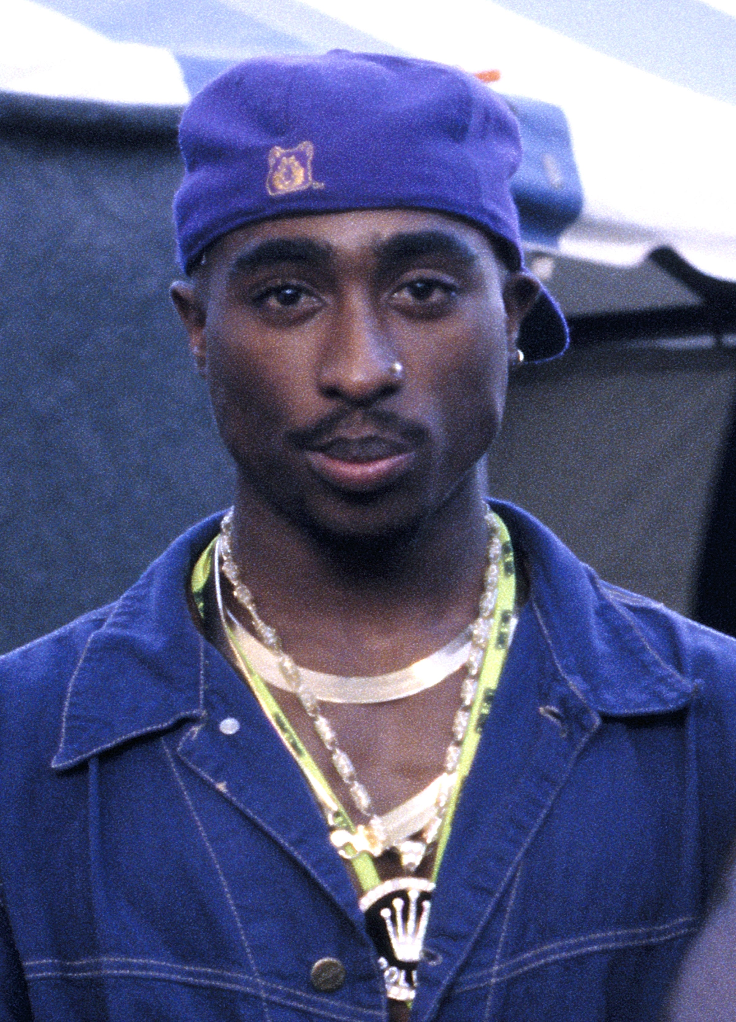 Tupac Shakur wearing a purple hat and layered necklaces. They have a focused expression and are dressed in a casual jacket
