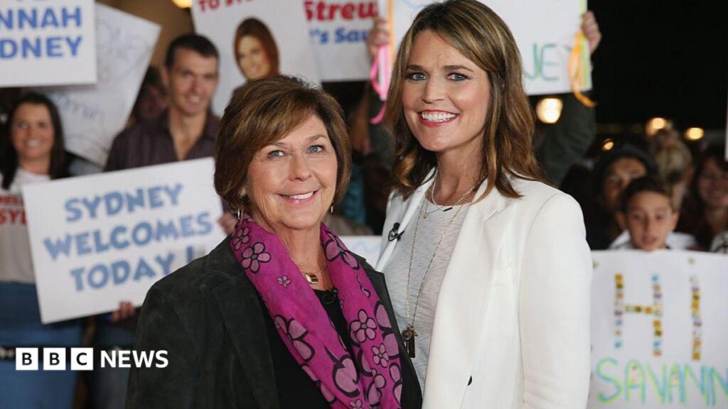 A woman with short brown hair looks directly into the camera with a slight smiling expression. She is wearing a pink top and a silver necklace with a heart charm.