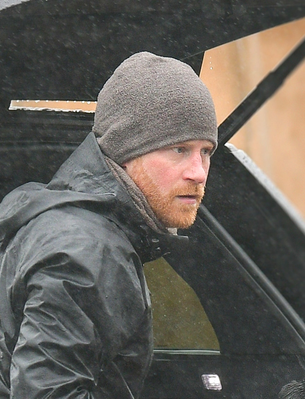 Prince Harry in a grey jacket and matching beanie standing in front of a car in Montecito looking serious.