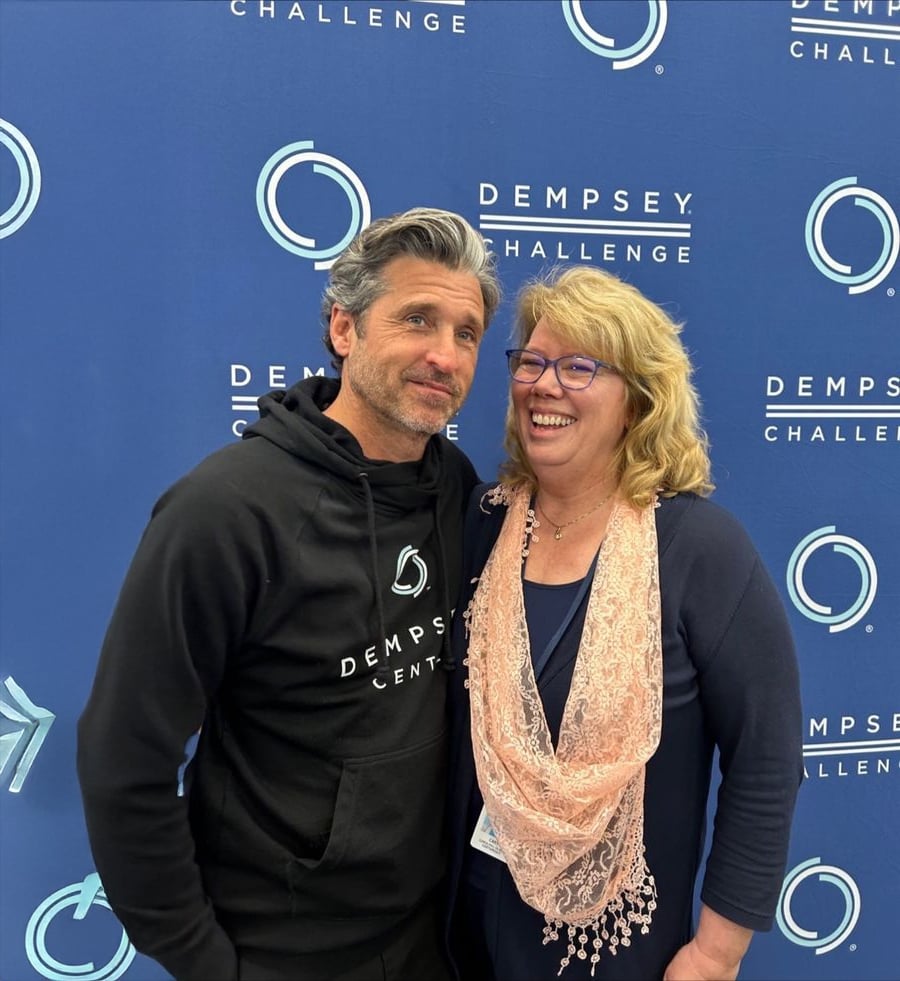 A man in a black "Dempsey Center" hoodie stands next to a smiling woman in glasses and a pink scarf, in front of a "Dempsey Challenge" backdrop.