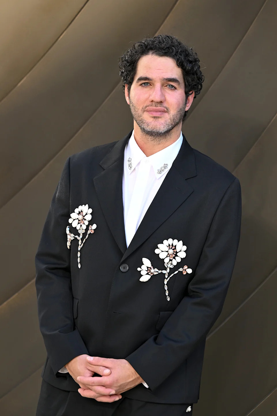 Person posing on a red carpet in a tailored black suit with floral embellishments on the jacket