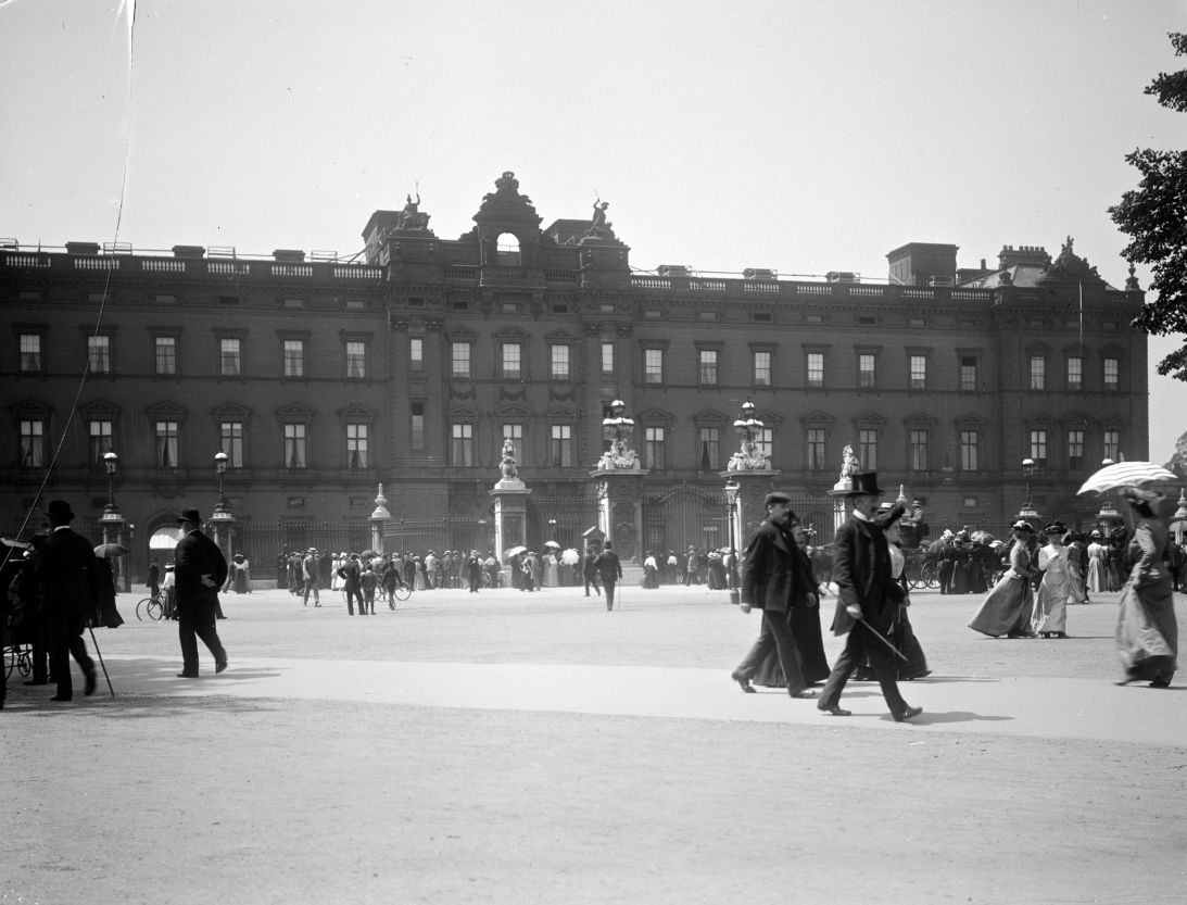 Sightseers crowd around the gates of Buckingham Palace, London. This view shows the public east facade as it looked after Edward Blore closed off the courtyard with a new wing in 1847 but before Aston Webb's more familiar refacing of 1913. The picture pre-dates the Queen Victoria Memorial, which was erected in 1911.