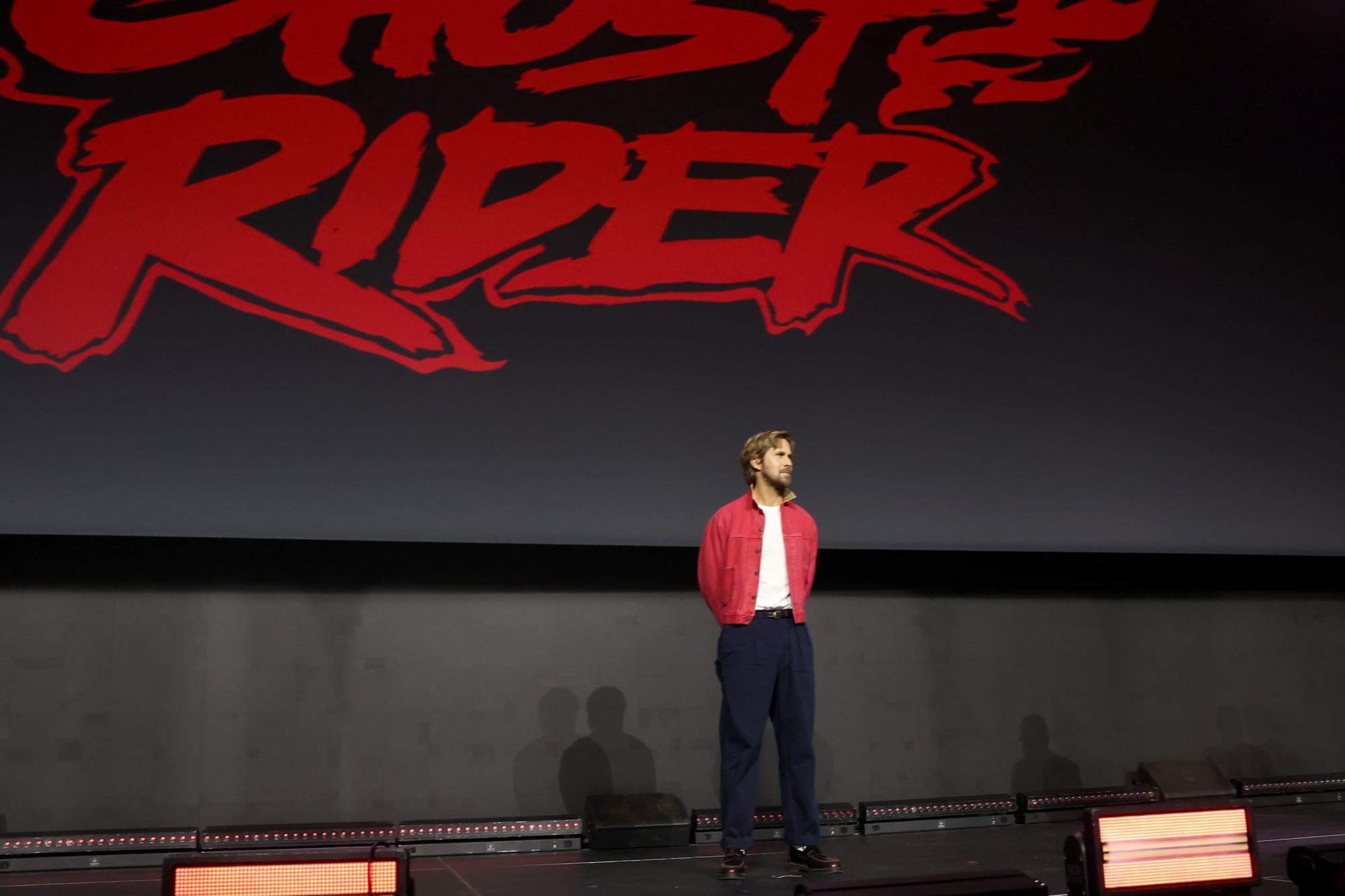 Ryan Gosling in red shirt standing under Ghost Rider logo