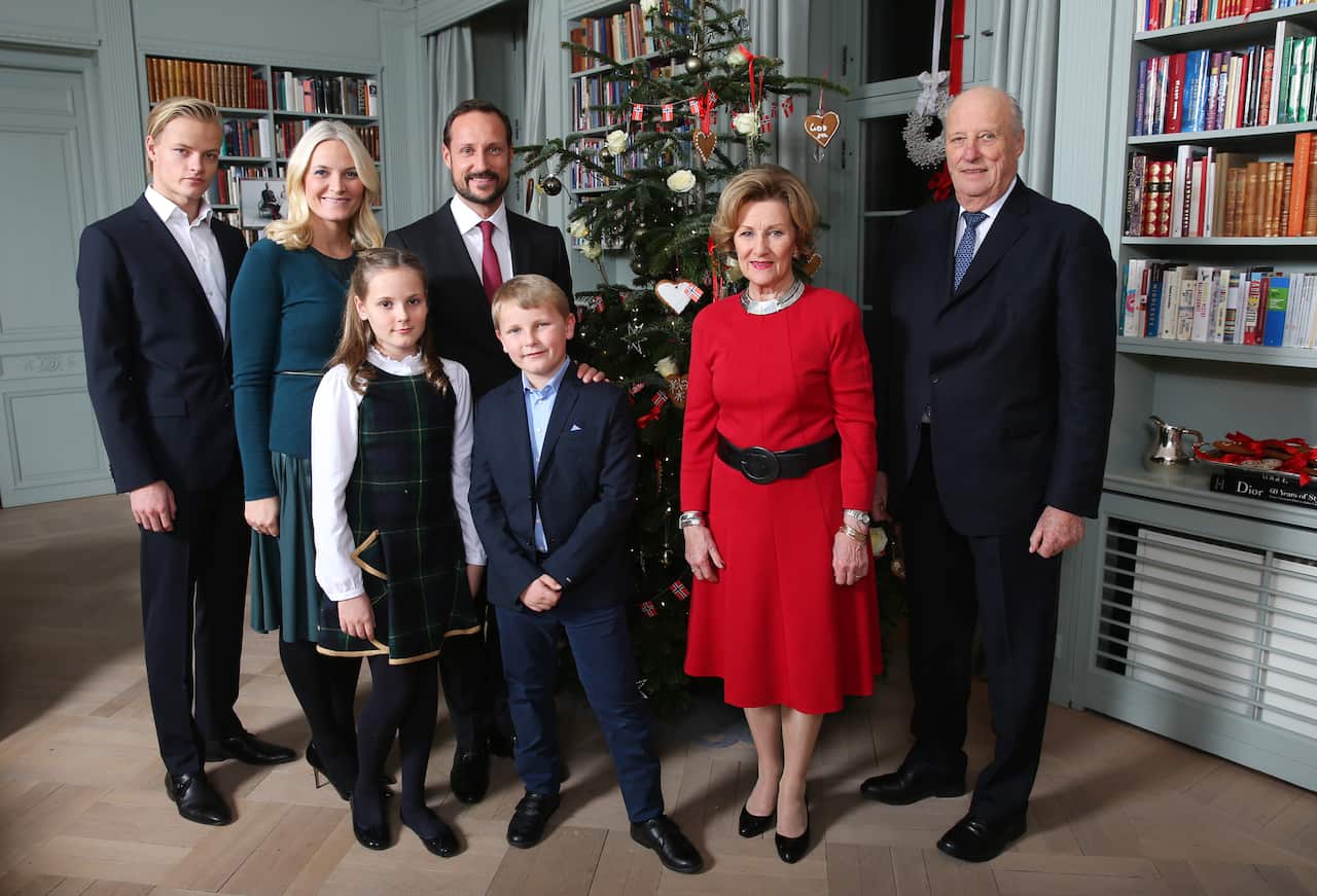 The Norwegian royal family stand in from of a Christmas tree. The Queen wears a bright red dress as children with blonde hair stand beside her.