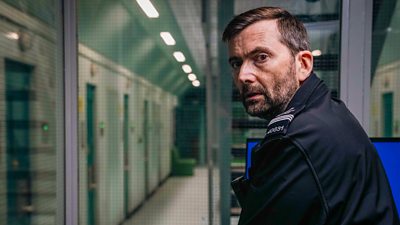 David Tennant in character as a uniformed Custodial Manager at Young Offenders Institution. Sitting in a glass-walled office, he looks over his shoulder at something offscreen.