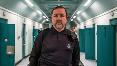 Photo of Daniel Ryan as Freddie Jennings in Time series 3. Wearing prison uniform, he walks down a prison corridor in between the open doors of prison cells.