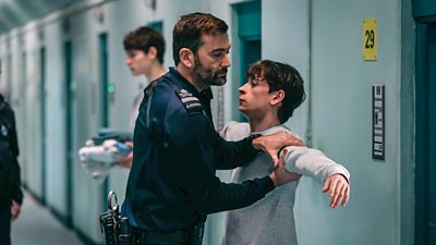 Bobby Bailey (David Tennant) pats the arm of James (Louis McCartney) as he checks for contraband. Bobby is dressed in prison uniform, while James wears a grey tracksuit. They stand in the corridor outside a prison cell.