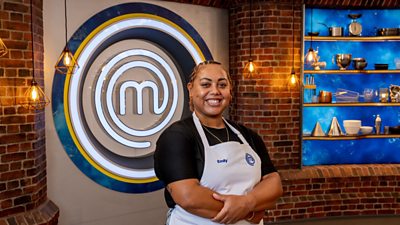 A woman in a black t-shirt and white apron  stood in the masterchef studio smiling towards camera