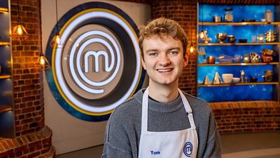 A man in a grey knitted jumper and a white apron stands smiling at the camera in the masterchef studio
