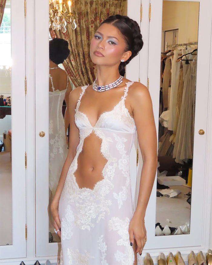 Zendaya modeling a white lace dress with cutouts and a statement necklace, reflecting her latest look in a mirror.