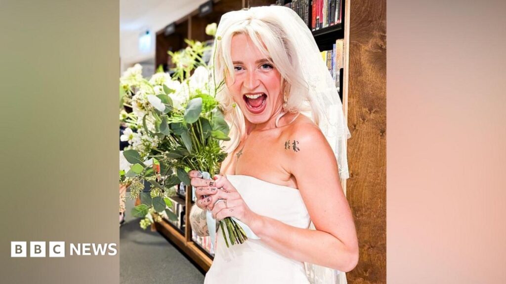 Bride dazzles in thrifty Chester wedding with £2 charity shop dress A woman with long blonde hair and wearing a white strapless wedding dress and white veil is smiling while holding a bouquet of flowers. She is standing in front of shelves of books as part of the Storyhouse library.