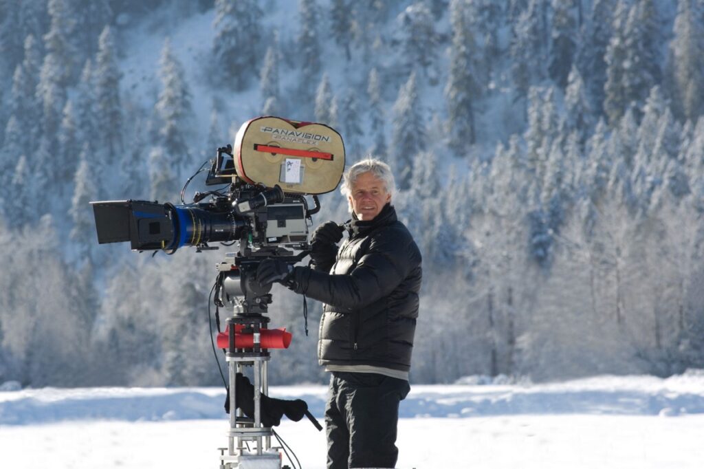 gray haired man in winter gear but bare-headed standing in snowy setting behind a movie camera